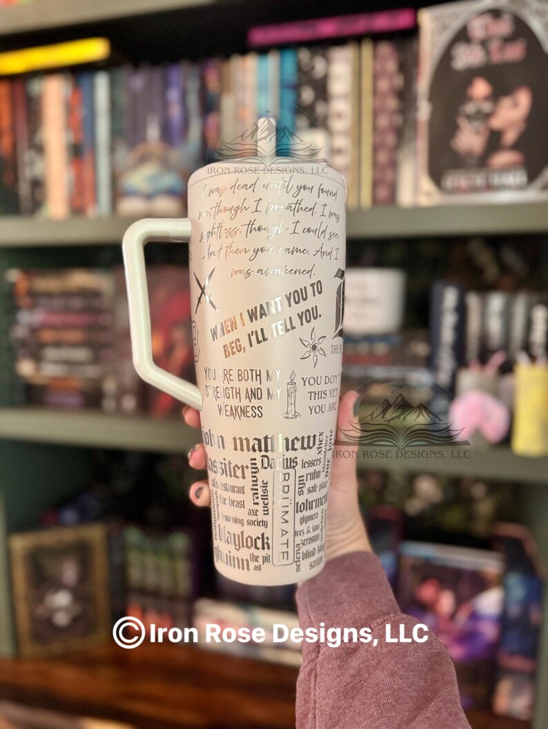 May include: A white tumbler with a large handle, featuring black text and graphics. The tumbler has quotes and words like "When I want you to beg, I'll tell you." and "John Matthew". The background is a bookshelf.