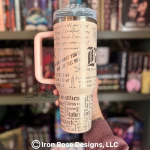 May include: A cream-colored tumbler with a light pink handle, featuring black text and graphics. The design includes quotes, a stylized "B", and the words "Matthew", "Darius", and "Kaylock". The tumbler has a silver lid with a mountain design.