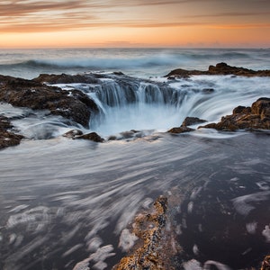 May include: A long exposure photograph of a natural rock formation with a large hole in the center. The hole is filled with water that is flowing in a circular motion. The water is a light blue color and the rocks are a dark brown color. The sky is a light orange color and the sun is setting in the distance.