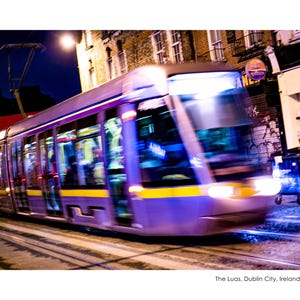 Könnte beinhalten: Ein sich schnell bewegendes lila-gelbes Straßenbahngleis bei Nacht. Die Scheinwerfer der Straßenbahn sind eingeschaltet, und das Bild ist verschwommen, was Bewegung suggeriert. Der Hintergrund umfasst Gebäude und Straßenlaternen. Der Text unten lautet "The Luas, Dublin City, Ireland."