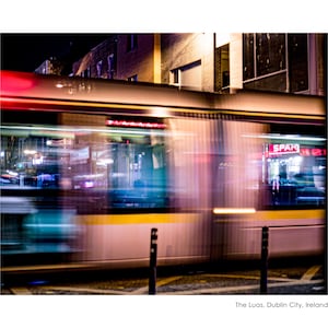 Könnte beinhalten: Langzeitbelichtungsfoto eines sich bewegenden Trams in Dublin City, Irland, bei Nacht. Das Tram ist weiß und gelb mit roten und blauen Lichtern, was einen Unschärfeeffekt erzeugt. Das SPAR-Schild ist sichtbar. Der Text lautet: The Luas, Dublin City, Ireland.