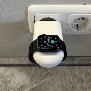 May include: A black Apple Watch with a black band is charging on a white wireless charging stand plugged into a wall socket.