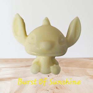 Puede incluir: Figura de color amarillo claro de un personaje de dibujos animados con orejas grandes, una nariz pequeña y una cabeza puntiaguda. La figura está sobre una superficie de madera. El texto "Burst Of Sunshine" está escrito en amarillo en la parte inferior.