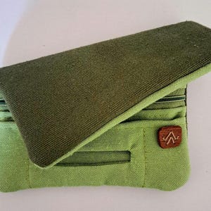 Op de afbeelding: Een groene canvas portemonnee met een flap, met een bruine leren patch met een bergachtig logo. De portemonnee heeft een vak met ritssluiting en meerdere kaartsleuven. De flap heeft een donkerdere groentint.