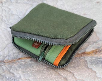 May include: An olive green canvas wallet with a gray zipper and a smaller, lighter green zippered compartment inside. The interior compartment has an orange stripe and a brown leather tag.