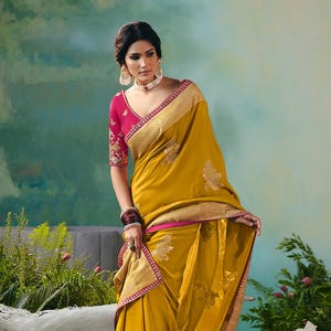 Może przedstawiać: Musztardowe sari ze złotym haftem kwiatowym i różową obwódką. Sari jest połączone z różową bluzką z haftowanymi rękawami. Kobieta ma na sobie biżuterię i stoi przed pawiem.