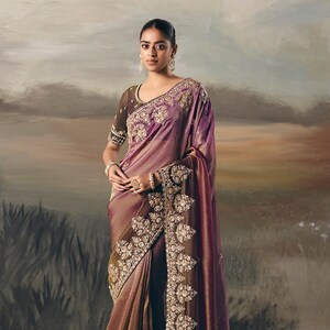 以下が含まれることがあります： 茶色と紫の色合いのフォーマルなサリーで、縁と胴には複雑な金の刺繍が施されています。 サリーは光沢のある質感で、お揃いのブラウスが付いています。 全体的なデザインはエレガントで、特別な機会に適しています。