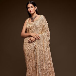 Puede incluir: Un sari de lentejuelas doradas con un delicado ribete de encaje. El sari está drapeado sobre una modelo y tiene un brillo sutil.
