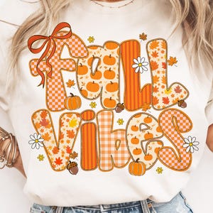 Pode incluir: Camiseta creme com as palavras "FALL VIBES" em laranja, branco e castanho. As letras são decoradas com abóboras, folhas e flores. A letra "F" tem um laço laranja.
