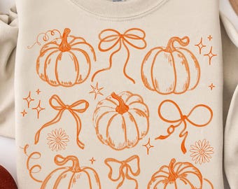 May include: Cream-colored sweatshirt featuring an orange pumpkin and bow design. The design includes various pumpkin illustrations, bows, stars, and floral accents. The sweatshirt is folded neatly, showcasing the fall-themed graphic.