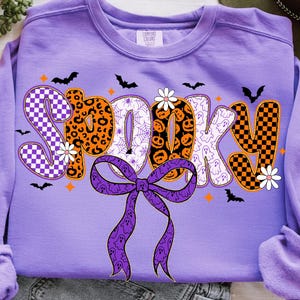 Może przedstawiać: Lawendowa bluza z napisem "Spooky" w różnych wzorach i wzorach o tematyce Halloween. Litery zawierają wzory w kratę, lamparcie, pajęczyny i duchy. Fioletowa kokarda i nietoperze dopełniają projekt.