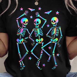 May include: Black t-shirt featuring a vibrant, rainbow-colored design of three dancing skeletons. The skeletons are surrounded by stars and bats. The design is eye-catching and perfect for Halloween or anyone who loves unique apparel.