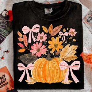 May include: Black crew neck sweatshirt featuring a pumpkin with a floral arrangement and pink bows. The flowers are in shades of pink and orange, with brown and orange leaves. A fall-themed design.