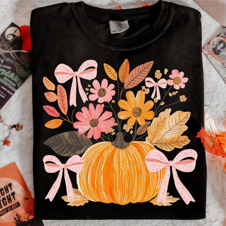 May include: Black crew neck sweatshirt featuring a pumpkin with a floral arrangement and pink bows. The flowers are in shades of pink and orange, with brown and orange leaves. A fall-themed design.