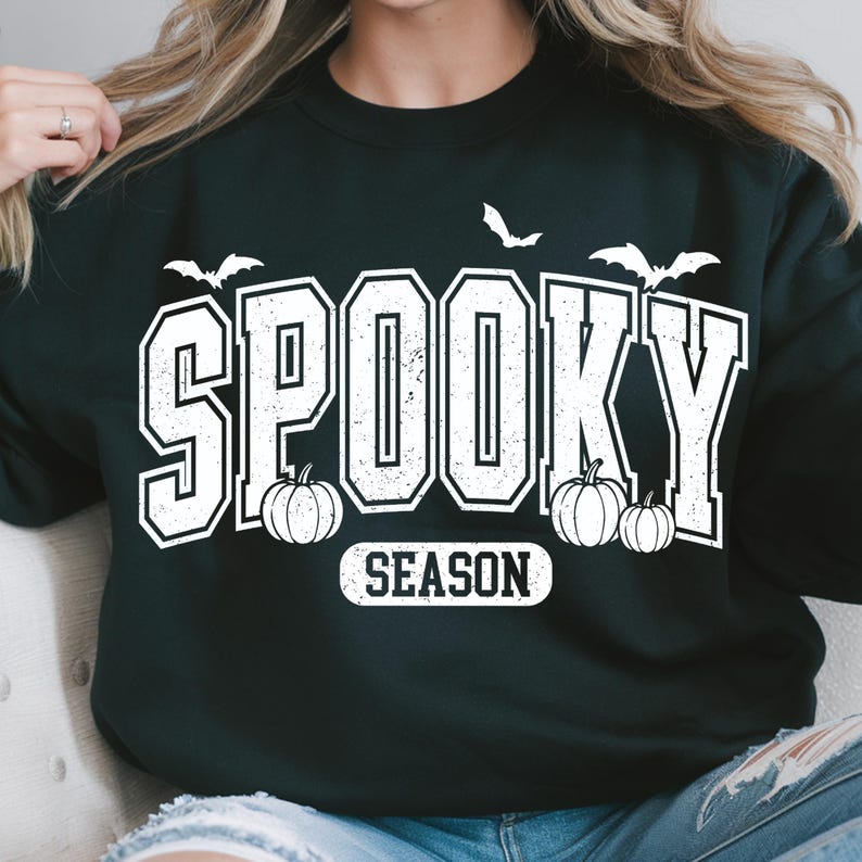 May include: Black sweatshirt with the words "SPOOKY SEASON" in white block letters. The design includes white bats and pumpkins, perfect for Halloween. A comfortable, casual top for the spooky season.