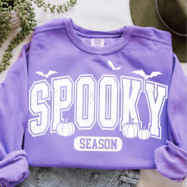 May include: A lilac-colored sweatshirt with the words "SPOOKY SEASON" in white block letters. The design includes white bats and pumpkins, perfect for Halloween. The sweatshirt is made of a soft material.