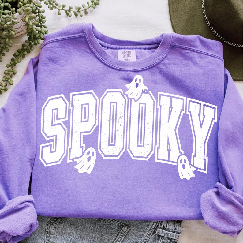 May include: A lilac-coloured sweatshirt featuring the word "SPOOKY" in large, distressed white lettering. Small white ghost graphics are scattered around the text. The sweatshirt is folded on a light-coloured surface.