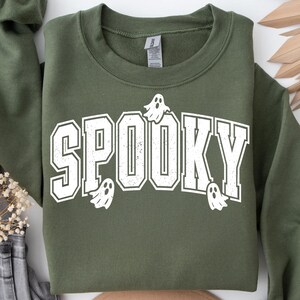 May include: An olive green sweatshirt featuring the word "SPOOKY" in large, white, block letters. The word "SPOOKY" has a distressed, vintage appearance. Small white ghost graphics are placed around the word. A crew neck sweatshirt.