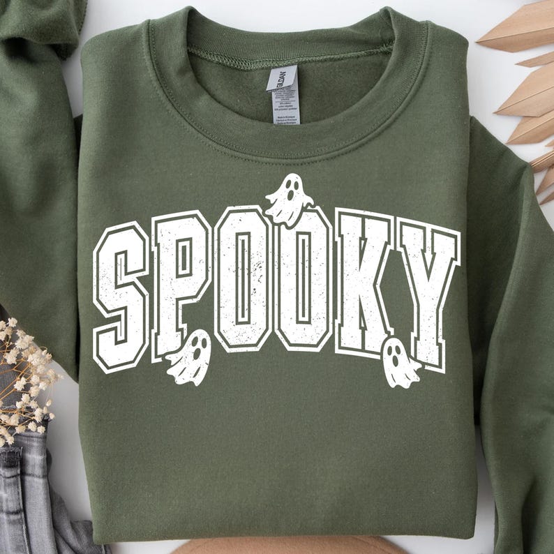 May include: An olive green sweatshirt featuring the word "SPOOKY" in large, white, block letters. The word "SPOOKY" has a distressed, vintage appearance. Small white ghost graphics are placed around the word. A crew neck sweatshirt.