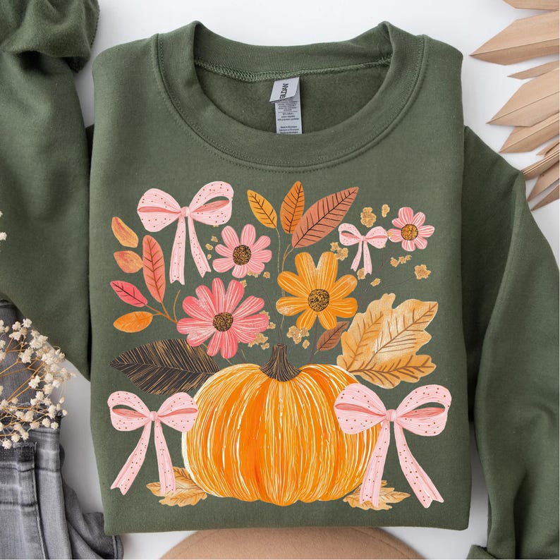 May include: An olive green sweatshirt featuring a fall-themed design. The graphic includes a large orange pumpkin, pink flowers, and autumn leaves. Pink bows are scattered around the floral arrangement. The sweatshirt has a crew neck and long sleeves.