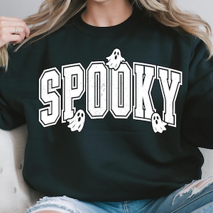 May include: Black sweatshirt featuring the word "SPOOKY" in bold, white lettering with a collegiate font. Small white ghost illustrations are placed around the text. A Halloween-themed top.