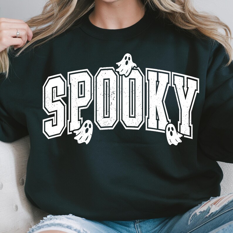 May include: Black sweatshirt featuring the word "SPOOKY" in bold, white lettering with a collegiate font. Small white ghost illustrations are placed around the text. A Halloween-themed top.