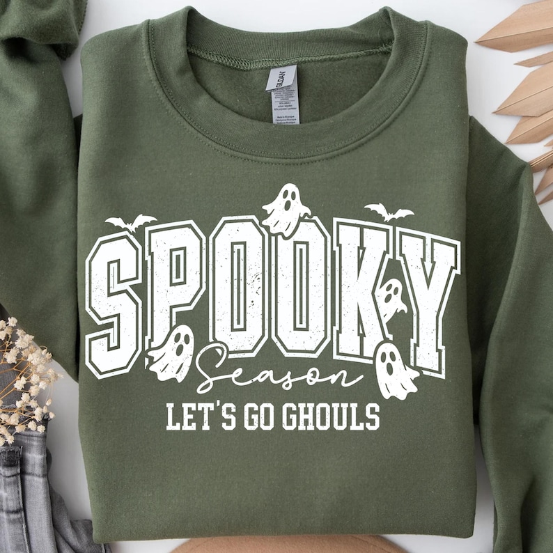 May include: Olive green sweatshirt with the words "SPOOKY SEASON" in white block letters, along with ghost and bat graphics. The phrase "LET'S GO GHOULS" is below the main text.