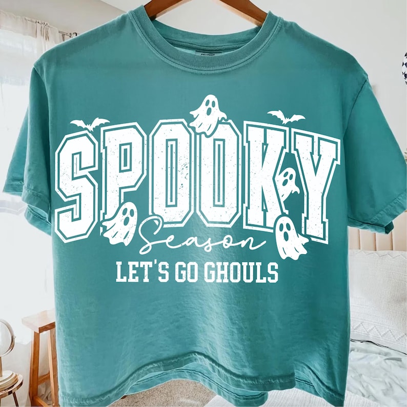 May include: Teal-colored t-shirt with the words "SPOOKY SEASON" in white block letters, accompanied by ghost and bat graphics. The phrase "LET'S GO GHOULS" is printed below. A casual, festive garment.