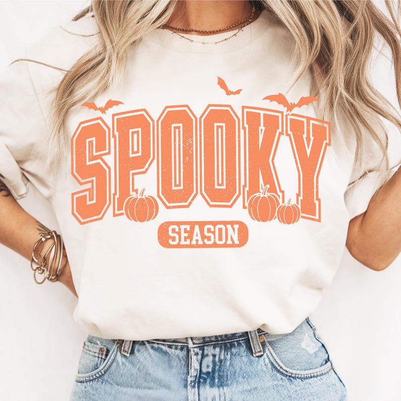 May include: Cream-colored t-shirt with the words "SPOOKY SEASON" in orange block letters. The design includes orange pumpkins and bats, evoking a Halloween theme. The shirt is a casual, comfortable style.