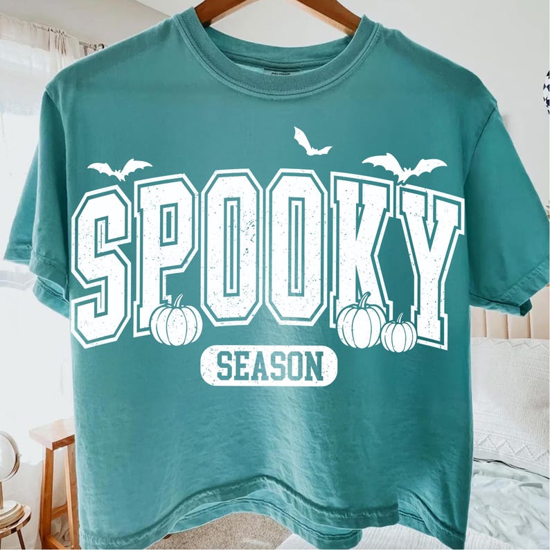 May include: Teal-colored t-shirt with the words "SPOOKY SEASON" in white block letters. The design includes white bats and pumpkins, perfect for Halloween. The shirt is a casual, short-sleeved style.