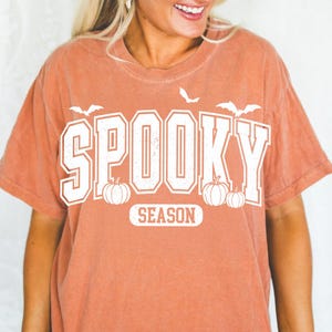 May include: A burnt orange t-shirt with the words "Spooky Season" in white block letters. The design includes white bats and pumpkins, perfect for Halloween. The shirt has a vintage, slightly faded look.