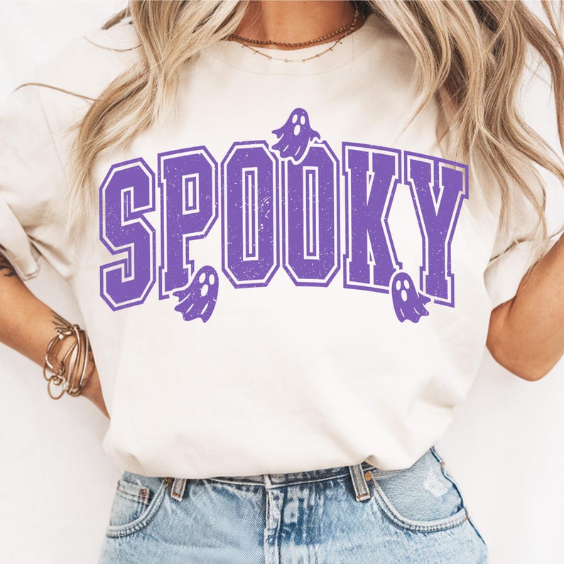May include: A cream-coloured t-shirt featuring the word "SPOOKY" in purple block letters, with three small ghost graphics. The shirt is a casual style, suitable for Halloween.