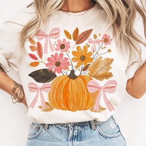 May include: Cream-colored t-shirt featuring a vibrant autumn design. The graphic includes an orange pumpkin, pink and orange flowers, brown leaves, and pink bows. A fall-themed tee for seasonal wear.