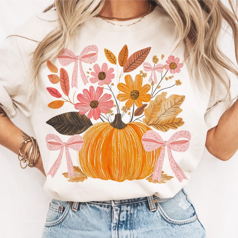 May include: Cream-colored t-shirt featuring a vibrant autumn design. The graphic includes an orange pumpkin, pink and orange flowers, brown leaves, and pink bows. A fall-themed tee for seasonal wear.