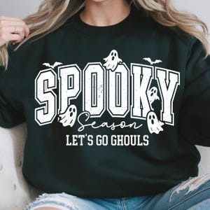 May include: Black sweatshirt with the words "Spooky Season" in white block letters, with ghost and bat graphics. The phrase "Let's Go Ghouls" is below the main text, also in white. A festive Halloween-themed design.