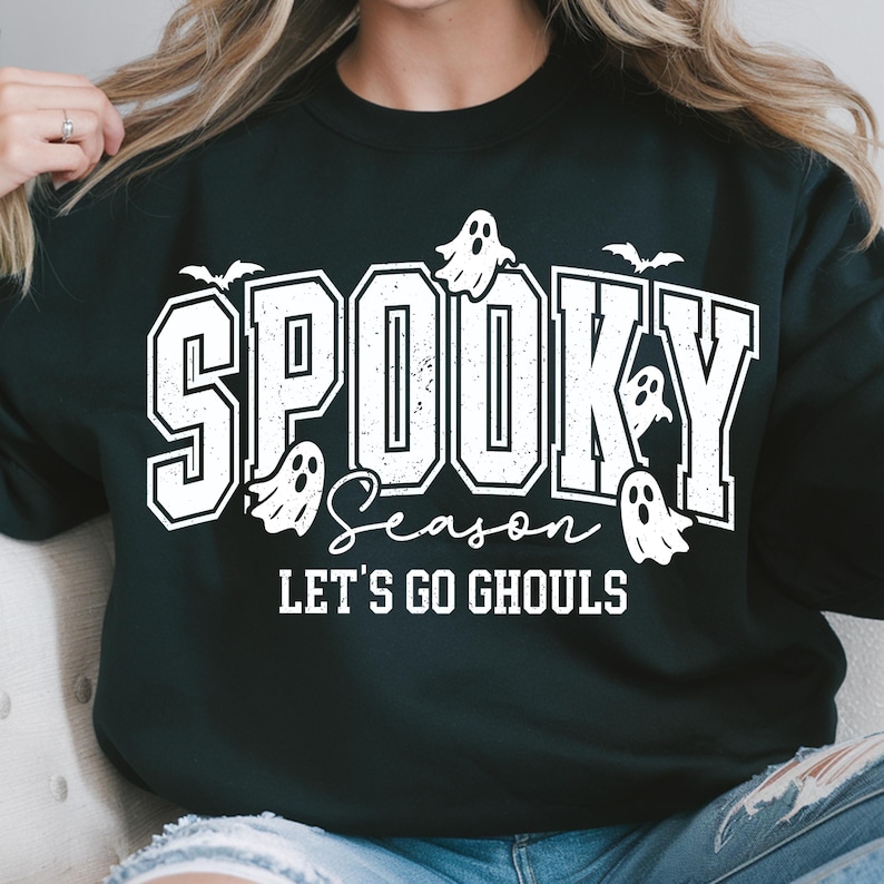 May include: Black sweatshirt with the words "Spooky Season" in white block letters, with ghost and bat graphics. The phrase "Let's Go Ghouls" is below the main text, also in white. A festive Halloween-themed design.