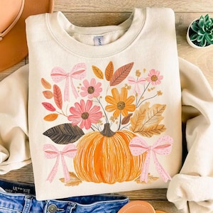 May include: Cream-colored sweatshirt featuring a vibrant fall-themed graphic. The design includes an orange pumpkin, pink and yellow flowers, brown leaves, and pink bows. A cozy and festive apparel item.