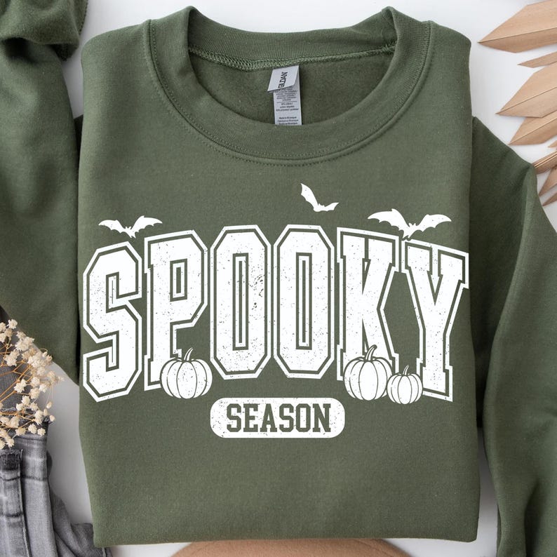 May include: An olive green sweatshirt with the words "SPOOKY SEASON" in white block letters. The design includes white bats and pumpkins, perfect for Halloween. The sweatshirt is a crew neck style.