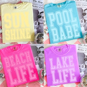 May include: Four colorful t-shirts with summer-themed text. The shirts are yellow, blue, pink, and purple. The yellow shirt says "SUN SHINE", the blue shirt says "POOL BABE", the pink shirt says "BEACH LIFE", and the purple shirt says "LAKE LIFE".