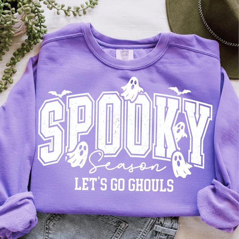 May include: A lavender sweatshirt with the words "Spooky Season" in white block letters, accompanied by ghost and bat graphics. The phrase "Let's Go Ghouls" is printed below. The sweatshirt is made of a soft material.