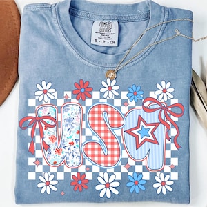 Könnte beinhalten: Hellblaues Comfort Colors T-Shirt mit dem Wort "USA" in einem patriotischen Design. Die Buchstaben zeigen Blumen-, Vichy- und Sternmuster mit roten Schleifen und Gänseblümchen-Akzenten. Eine goldene Halskette liegt auf dem Shirt.