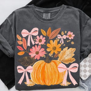May include: Dark gray sweatshirt featuring a vibrant autumn design. An orange pumpkin is surrounded by colorful flowers, leaves, and pink bows. The design evokes a fall harvest theme.