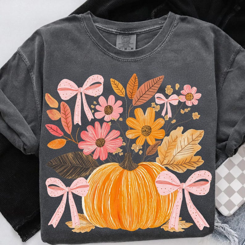 May include: Dark gray sweatshirt featuring a vibrant autumn design. An orange pumpkin is surrounded by colorful flowers, leaves, and pink bows. The design evokes a fall harvest theme.