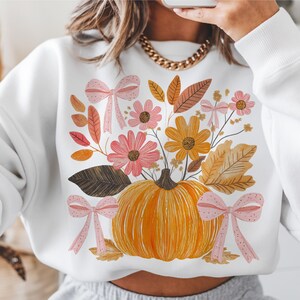 May include: White sweatshirt featuring a pumpkin with a floral arrangement of pink and yellow flowers, brown leaves, and pink bows. The pumpkin is orange, and the flowers and leaves have autumn colors. The design is centered on the front of the sweatshirt.
