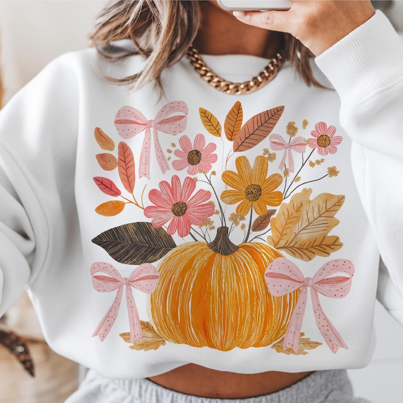 May include: White sweatshirt featuring a pumpkin with a floral arrangement of pink and yellow flowers, brown leaves, and pink bows. The pumpkin is orange, and the flowers and leaves have autumn colors. The design is centered on the front of the sweatshirt.