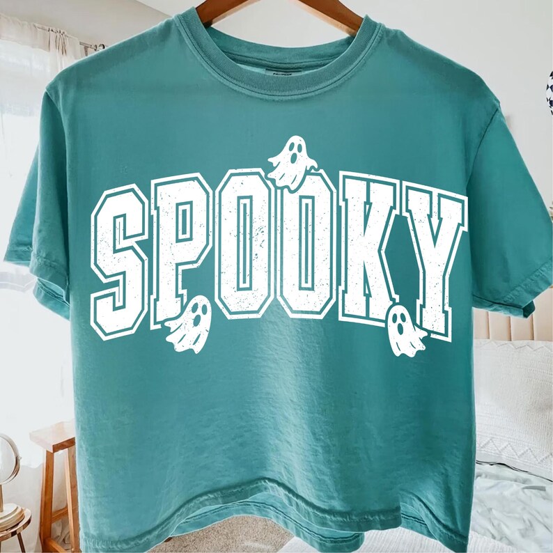 May include: Teal t-shirt featuring the word "SPOOKY" in large, white, collegiate-style lettering, with white ghost graphics. The t-shirt is displayed on a wooden hanger.