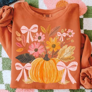 May include: Rust-colored sweatshirt featuring a floral and pumpkin design. The graphic includes a large orange pumpkin, pink and yellow flowers, and pink bows. The text "COMFORT COLORS" is visible on the upper right.