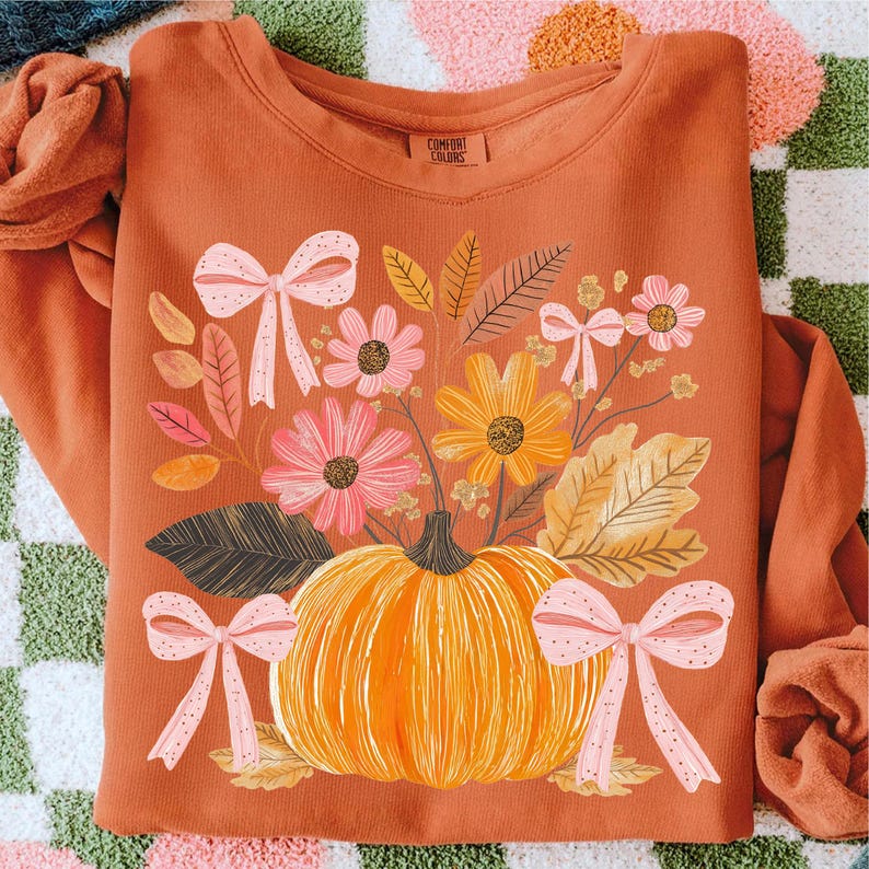 May include: Rust-colored sweatshirt featuring a floral and pumpkin design. The graphic includes a large orange pumpkin, pink and yellow flowers, and pink bows. The text "COMFORT COLORS" is visible on the upper right.