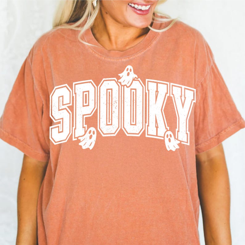 May include: A terracotta-coloured t-shirt featuring the word "SPOOKY" in large, white, collegiate-style lettering. Small white ghost graphics are scattered around the text. The shirt has a slightly faded, vintage appearance.
