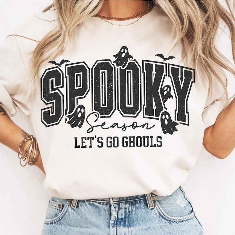 May include: A cream-colored t-shirt with the words "SPOOKY Season LET'S GO GHOULS" in black lettering. The design includes ghost and bat graphics, perfect for Halloween.